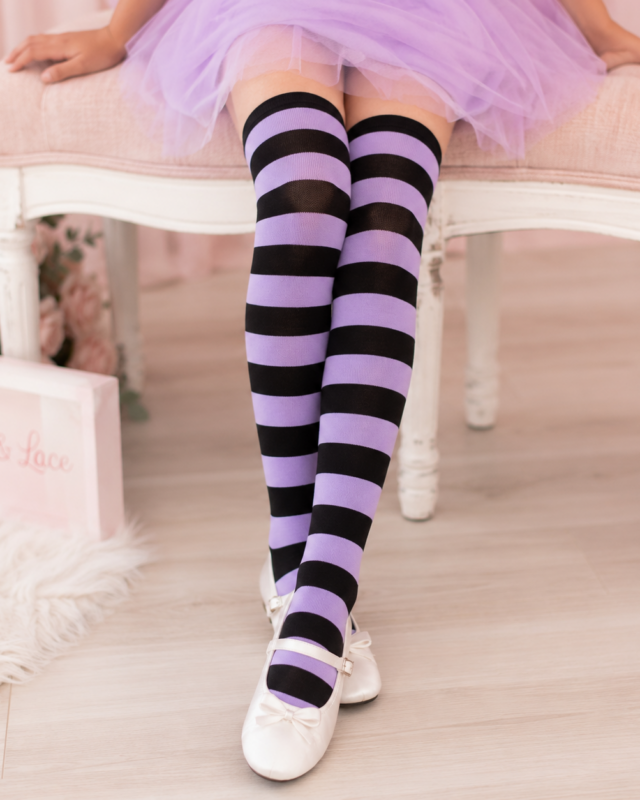 A person seated on a pink bench wearing lavender and black horizontally striped over-the-knee socks with white ballet flats. The setting is a softly lit indoor room with a pale wood floor, pink flowers, and a pink box labeled 'Helly & Lace' visible on the left.