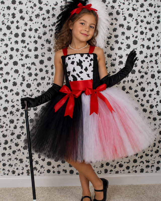 A young girl modeling a black and white cow-print costume dress with a red waist sash and bow, black gloves, a black feathered headband with white feathers, and a black-and-white split tutu skirt. She holds a cane and stands against a black and white patterned wall.