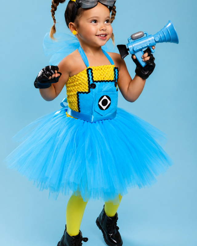 A young girl modeling a Minions costume against a solid light blue background. The costume features a bright blue tutu skirt, a yellow and blue vest with a Minions logo, yellow tights, black shoes, and a Minions headband with goggles.